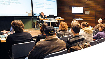 Mensch mit Behinderung hält einen Vortrag auf einer Bühne vor Publikum im Saal