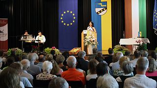 Aras steht am Rednerpult vor Publikum in einem Saal.