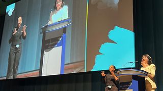 Aras spricht auf dem Podium. Eine Gebärdendolmetscherin übersetzt die Rede mit Gesten