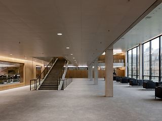 Moderne, offene Innenhalle mit einer zentralen Treppe, großen Glasfronten, Sitzbereichen aus schwarzen Sesseln und Blick auf ein historisches Gebäude im Außenbereich.