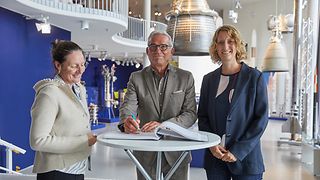 Landtagspräsident Strobl schreibt ins Gästebuch des DLR-Standorts. Daneben Dr. Anke Kovar vom DLR und die Abgeordnete Isabell Huber. Im Hintergrund eine Ausstellung zur Raumfahrt.