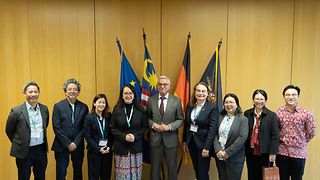 Landtagspräsident Strobl posiert mit acht Delegierten aus Malaysia vor den Länderflaggen. Strobl steht in der Mitte, im Hintergrund ist eine holzgetäfelte Wand.