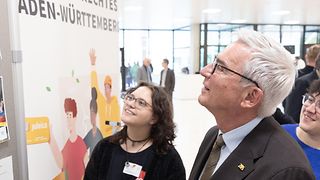 Landtagspräsident Strobl betrachtet gemeinsam mit zwei Frauen den Infostand