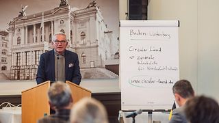 Präsident Strobl steht an einem Rednerpult in einem Konferenzsaal und spricht ins Mikro. Neben ihm steht ein Flipchart mit Informationen zum Projekt "Circular Länd".