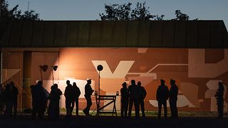 Das Foto zeigt das Gebäude, auf dessen Mauer das Gedenk-Graffiti angebracht wurde, bei Nacht. Das Graffiti wird von Scheinwerfern beleuchtet. Davor sind die Silhouetten der anwesenden Personen zu sehen.