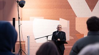 Landtagspräsident Strobl spricht an einem Rednerpult vor der durch Scheinwerfer beleuchteten Wand mit einem Graffiti. Am unteren Bildrand lassen sich die Umrisse der Zuhörenden erahnen.